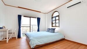 Une chambre avec un plancher en bois, un lit recouvert d’une courtepointe bleu clair, un bureau avec une chaise, une fenêtre avec des rideaux bleus et un appareil de climatisation.