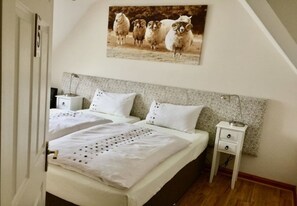 A bedroom with two beds, a headboard with a textured pattern, a wall-mounted picture of sheep, bedside tables with lamps, and a door with a number.