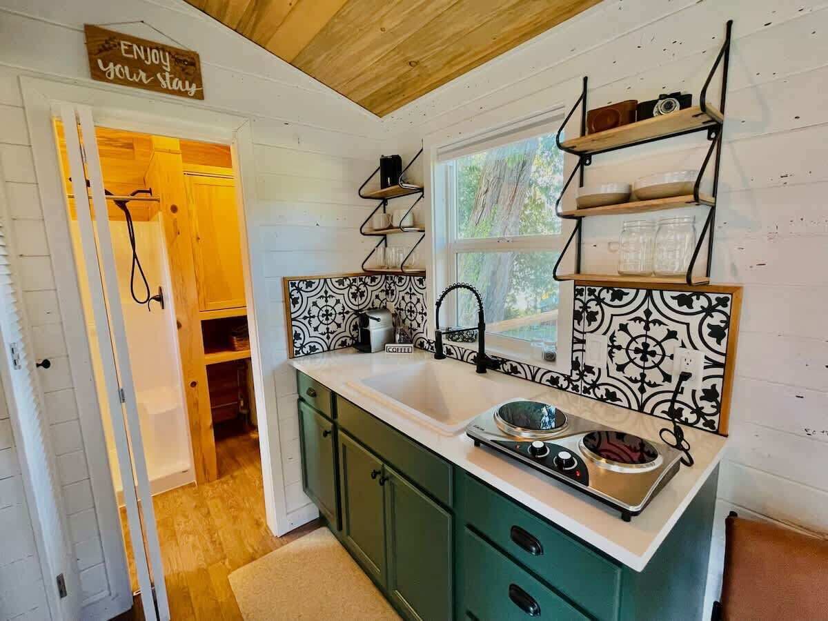A compact kitchen with a sink, stove, and green cabinets. A window provides natural light, and a sign above the door reads "Enjoy your stay."