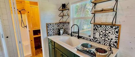 A compact kitchen with a sink, stove, and green cabinets. A window provides natural light, and a sign above the door reads "Enjoy your stay."