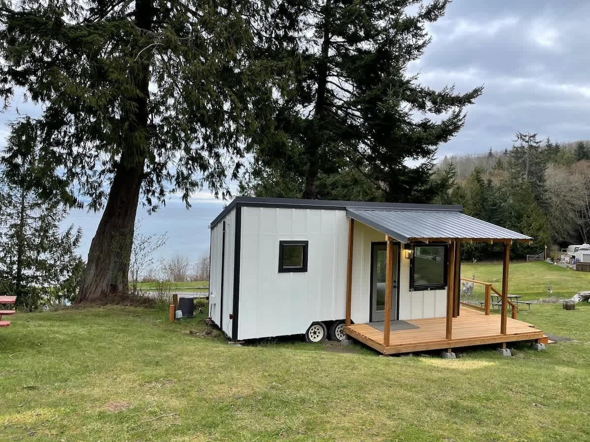 A small, single-story cabin with a porch and a small deck, situated on a grassy area near a body of water.