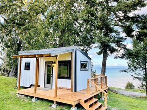 A small cabin with a deck, a door, and windows, situated on a grassy area with trees and a view of the water. - Moonlight Ridge Picturesque Tiny Home with a Milli (Port Angeles)