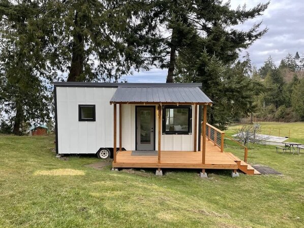 A small cabin with a wooden deck and a metal roof, situated in a grassy area with trees in the background. - Moonlight Ridge Picturesque Tiny Home with a Milli (Port Angeles)
