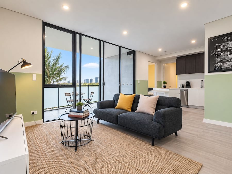 A modern living room with a sofa, a coffee table, and a TV. There is a large window offering a view of the outdoors.