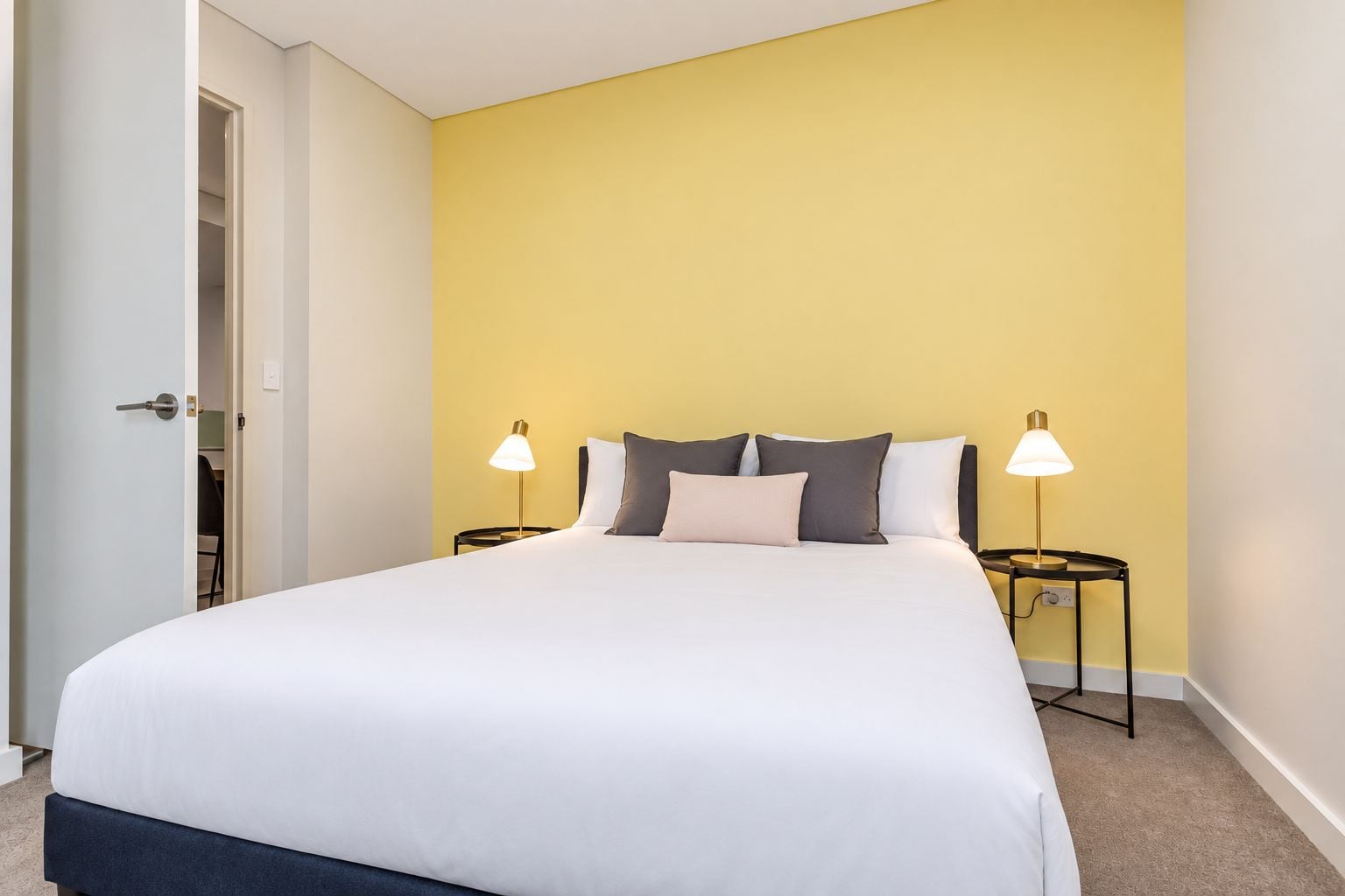 Una cama bien hecha con edredón blanco y varios almohadones, dos lámparas de noche y una puerta que da a un armario.
