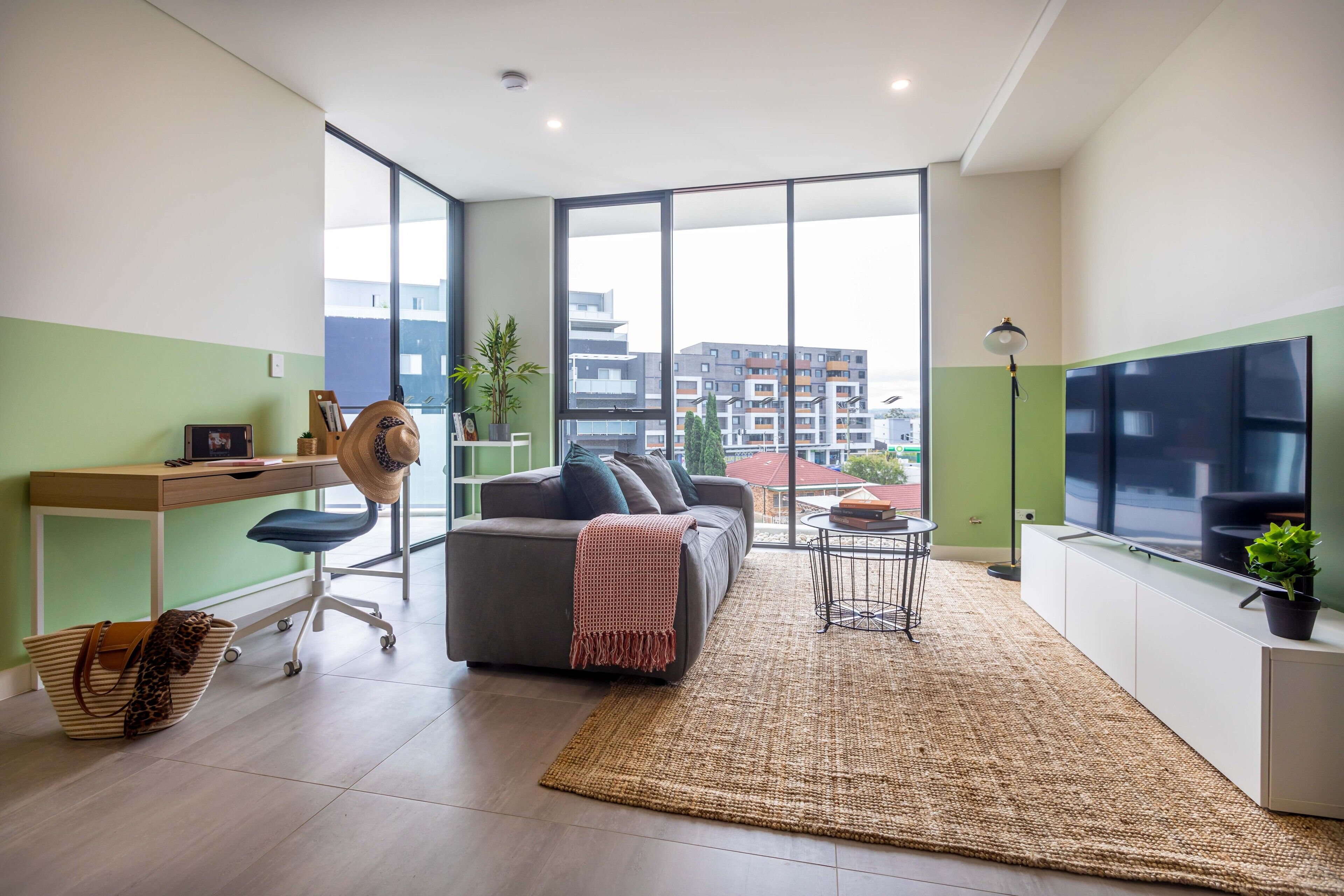 A modern living room with a sofa, desk, and large windows offering a city view.