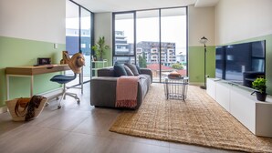 A modern living room with a sofa, desk, and large windows offering a city view. - KULA - Apartment Parramatta (Mays Hill)