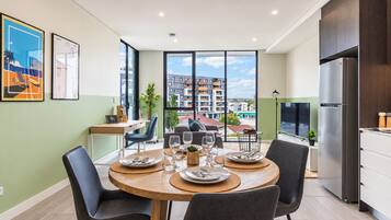 A modern dining area with a round table set for four, a kitchen with a stainless steel refrigerator, and a view of a cityscape through large windows.