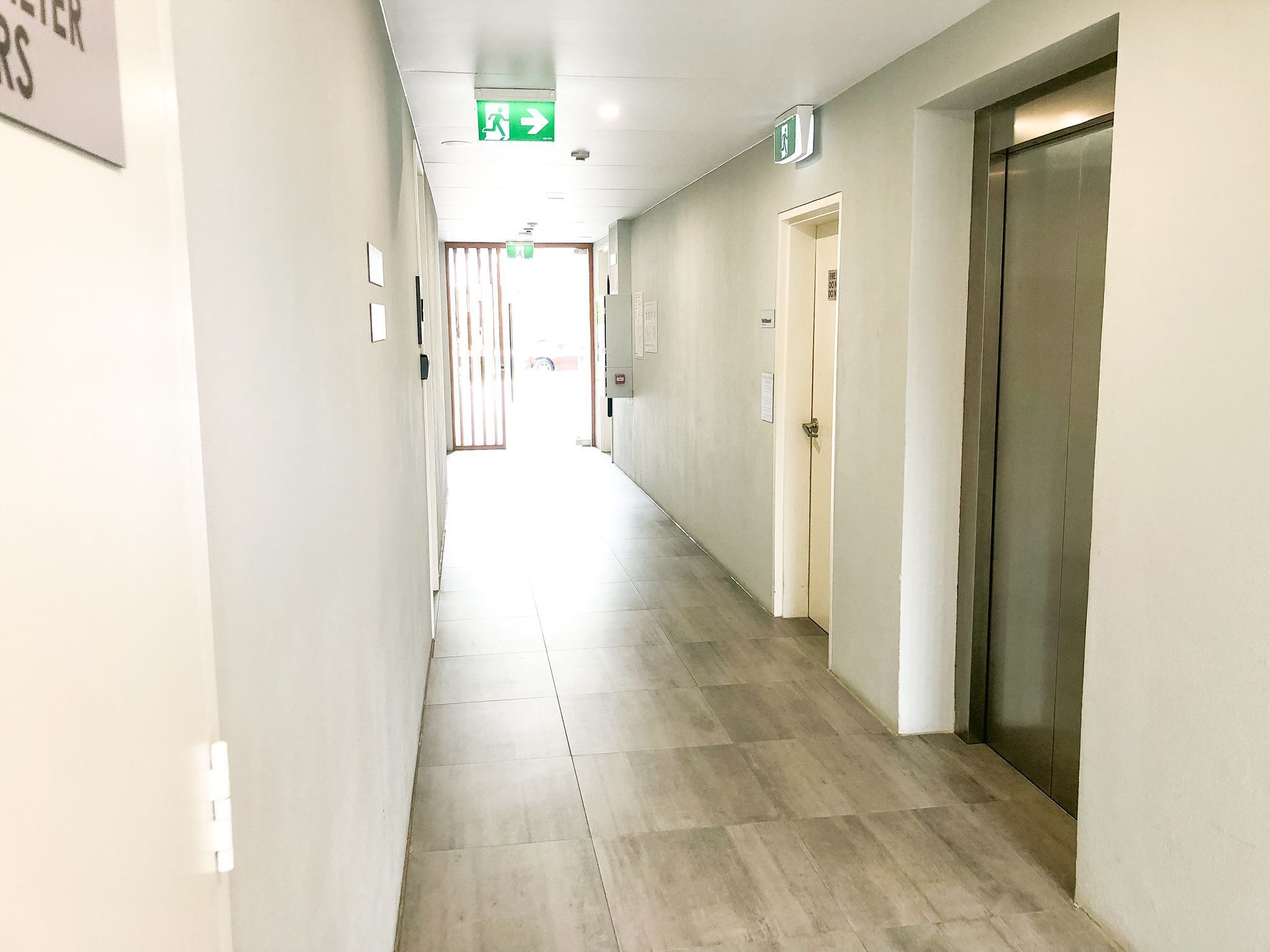 A long, narrow hallway with a tiled floor, doors on the right, and emergency exit signs on the ceiling.