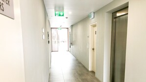 A long, narrow hallway with a tiled floor, doors on the right, and emergency exit signs on the ceiling. - KULA - Apartment Parramatta (Mays Hill)