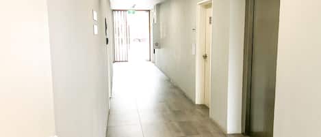 A long, narrow hallway with a tiled floor, doors on the right, and emergency exit signs on the ceiling.
