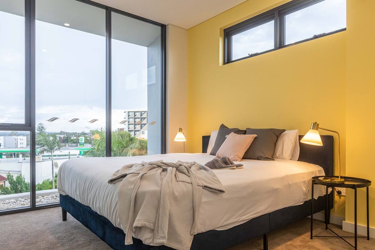Ein modernes Schlafzimmer mit einem großen Bett, einem Nachttisch und Blick auf die Stadt durch raumhohe Fenster.