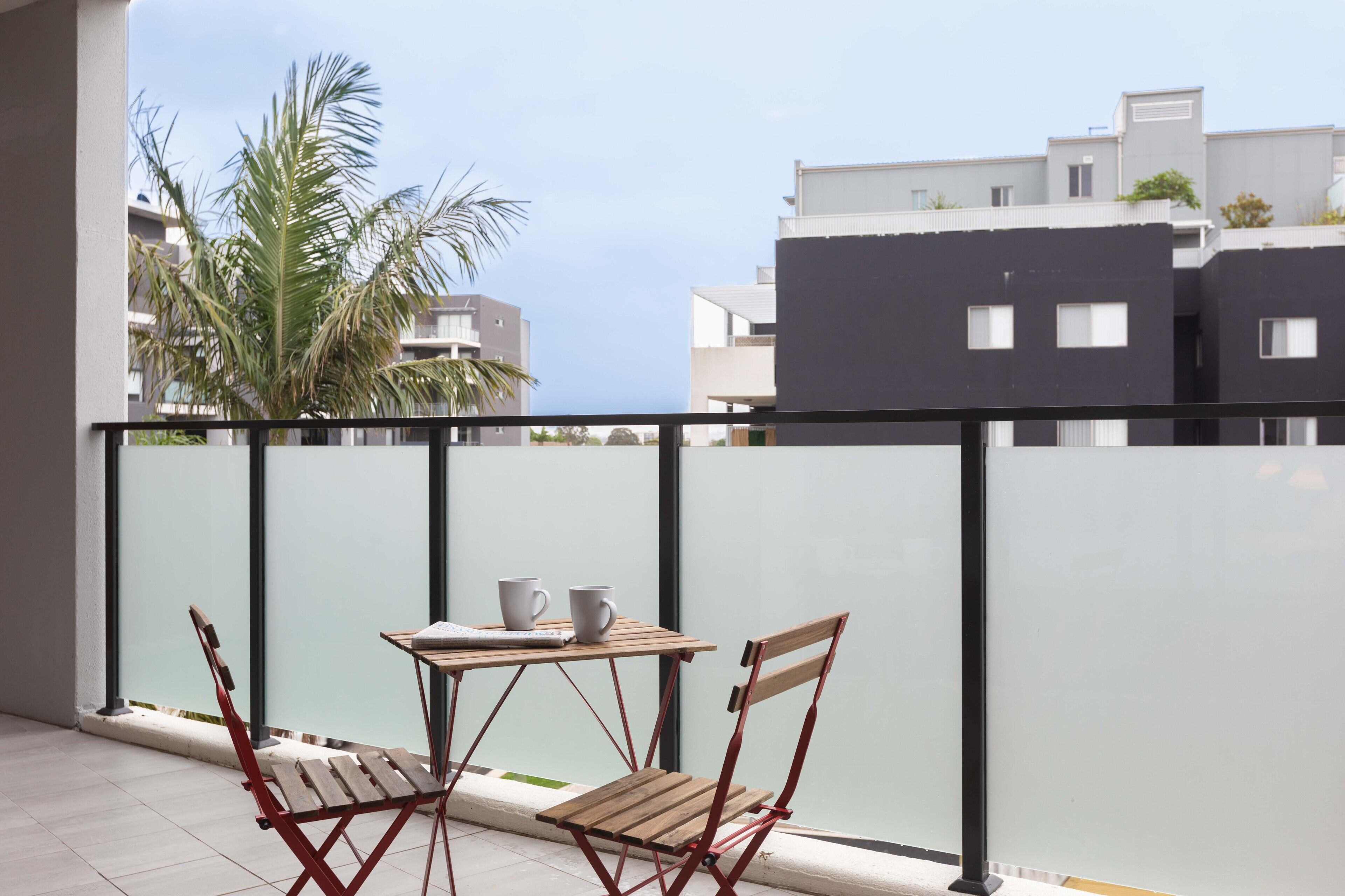 A balcony with a table and two chairs, overlooking a residential area with modern buildings and a palm tree.