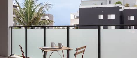 A balcony with a table and two chairs, overlooking a residential area with modern buildings and a palm tree.
