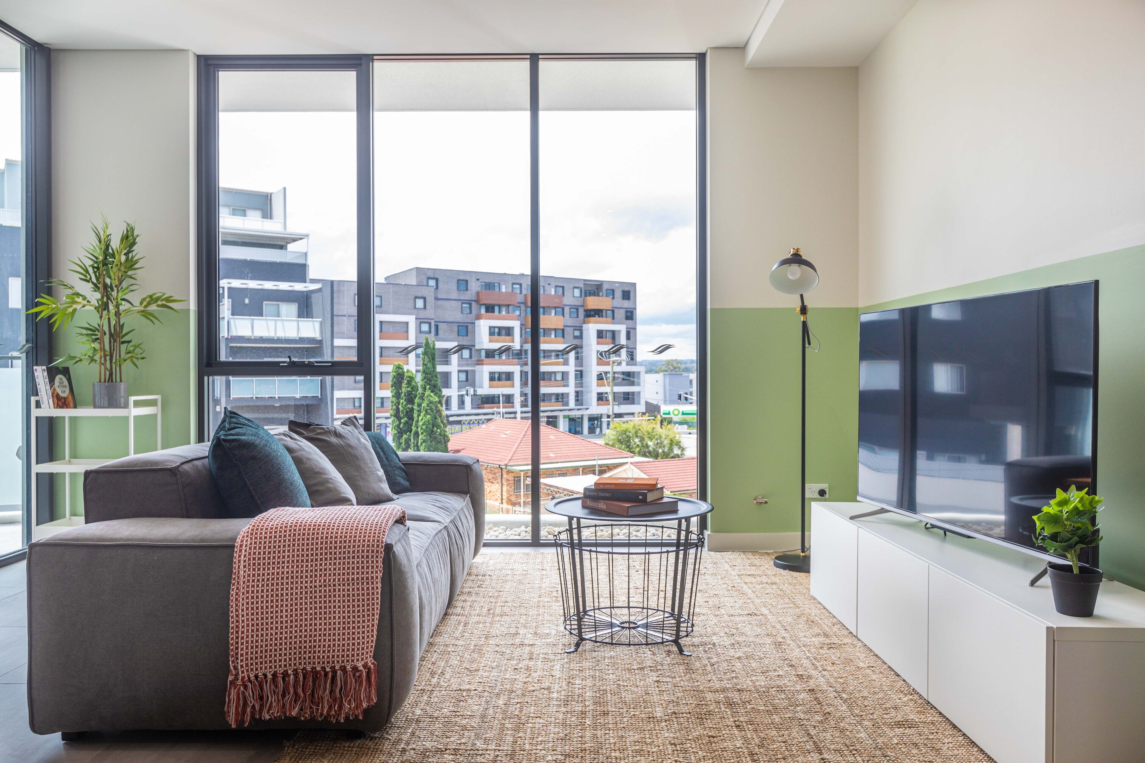 A modern living room with a sofa, a small round table, a floor lamp, and a large window offering a view of buildings.