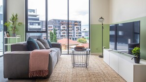 A modern living room with a sofa, a small round table, a floor lamp, and a large window offering a view of buildings. - KULA - Apartment Parramatta (Mays Hill)