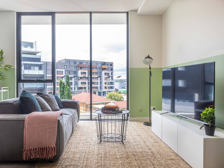 En moderne stue med sofa, rundt sofabord, gulvlampe og et stort vindue med udsigt til bygninger.