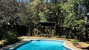 A clear, curved swimming pool with a pebble-lined edge, surrounded by a wooden fence and lush greenery.