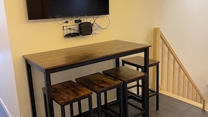 Un coin repas avec une table en bois et trois chaises, un téléviseur à écran plat fixé au mur, et un escalier avec des rampes en bois.