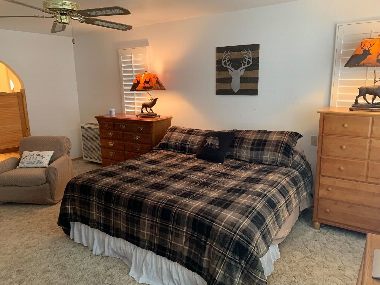 Une chambre avec une couvre-lit à carreaux, une commode en bois, une chaise, une lampe et une œuvre d’art murale.