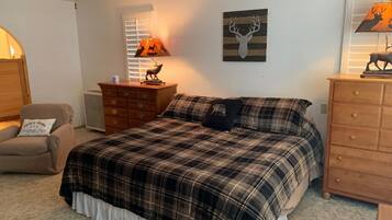 Une chambre avec une couvre-lit à carreaux, une commode en bois, une chaise, une lampe et une œuvre d’art murale.