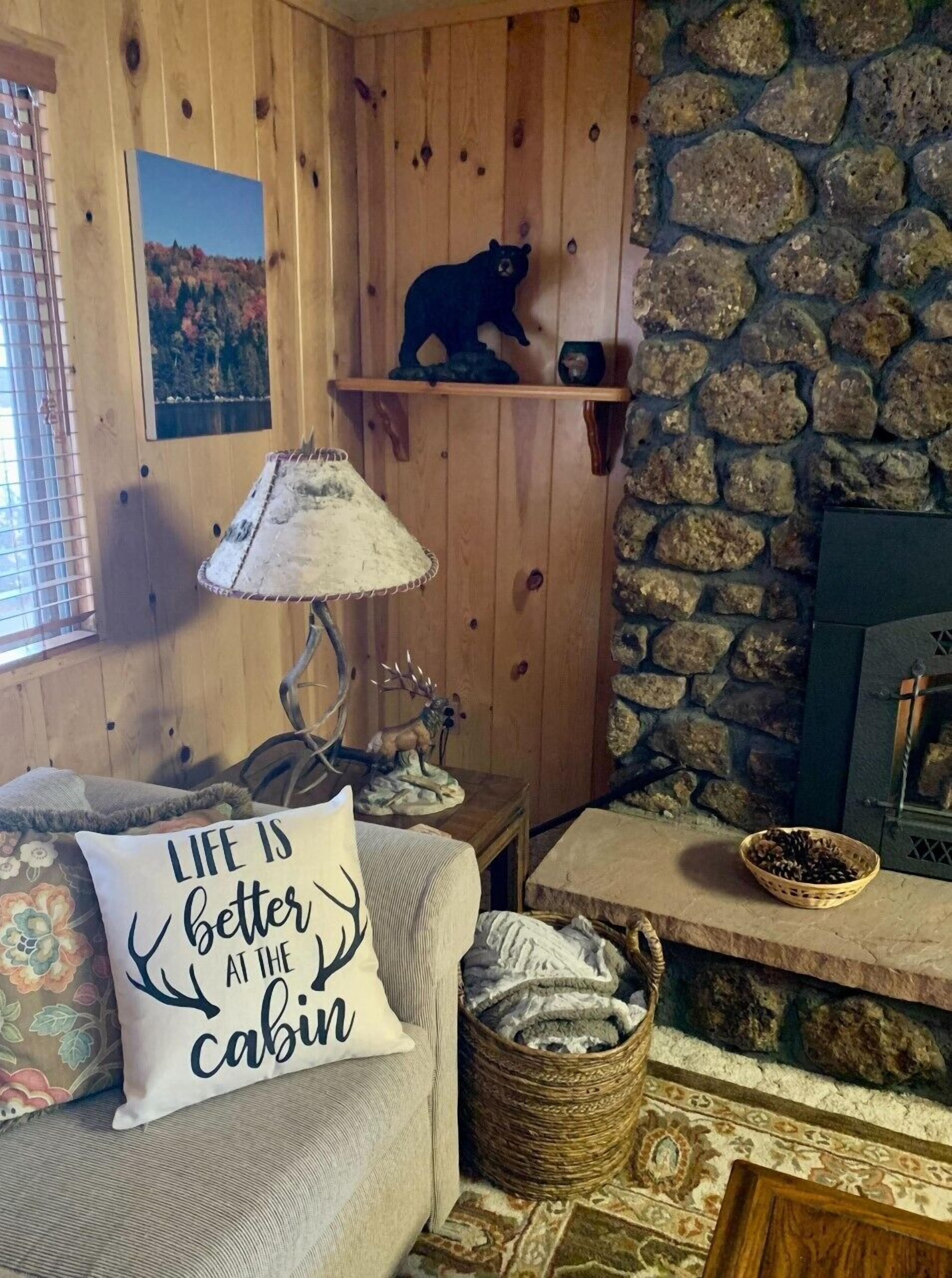 A cozy cabin interior with wooden walls, a stone fireplace, a bear decoration, a lamp, and a cushion with a cabin-themed message.