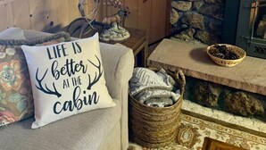 Un accogliente interno di una baita con pareti in legno, un camino in pietra, una decorazione a forma di orso, una lampada e un cuscino con un messaggio a tema baita.