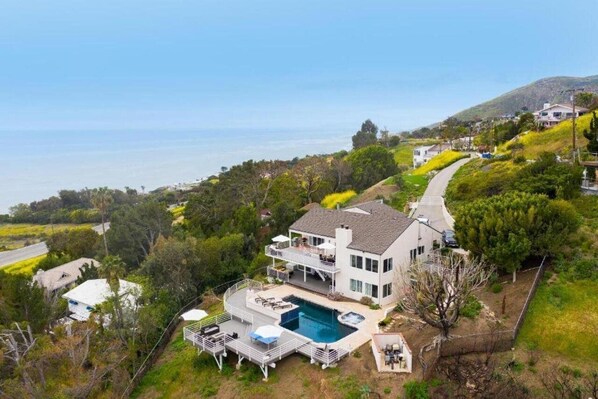 Une maison à deux étages avec une piscine, entourée de verdure et offrant une vue sur l’océan.