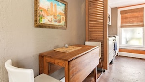 A kitchen with a wooden table, white chairs, a painting on the wall, and a window with blinds.