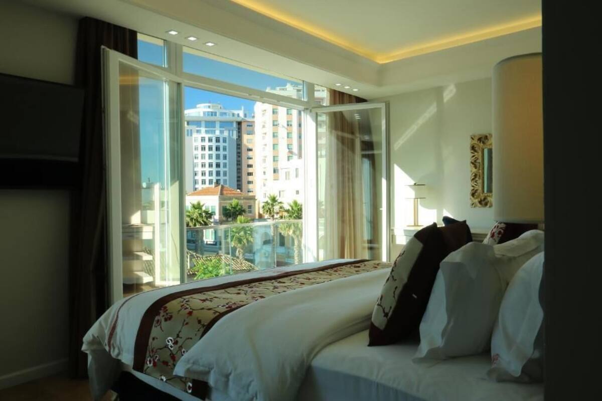 Ein Hotelzimmer mit einem großen Bett, Blick auf eine Stadtlandschaft durch eine Schiebetür und einem Balkon mit Pool.