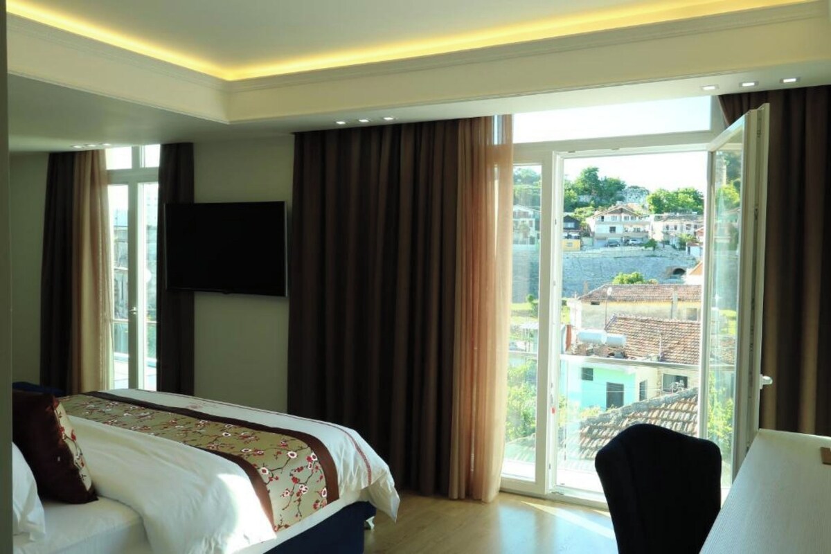 Habitación de hotel con cama, televisor, ventana grande con cortinas y vista a edificios.