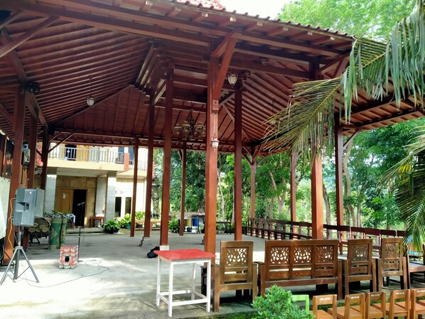 Un espacio exterior cubierto con mobiliario de madera y una mesa roja, rodeado de follaje y un edificio al fondo.