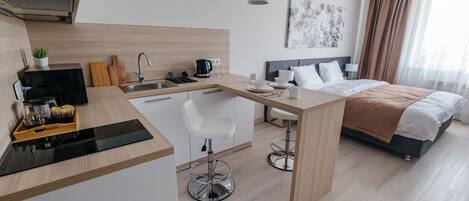 A modern hotel room with a kitchenette, a bed, a dining area, and a large window with curtains.