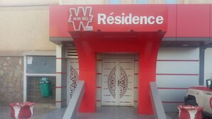 A hotel entrance with a red and white facade, a large sign reading "WIN BELY Résidence Hotel", and two symmetrical decorative pillars. - Winbel Hotel (Hassi Messaoud)