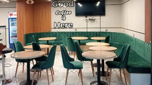 A modern coffee shop interior with green upholstered seating, round tables, and a sign that reads "Good Coffee is Here." - Winbel Hotel (Hassi Messaoud)