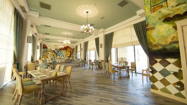 A spacious dining area with multiple tables and chairs, a chandelier, and large windows with curtains.