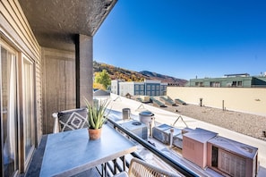 A balcony with a table, chairs, and a potted plant. There are air conditioning units and a view of buildings and hills in the background.