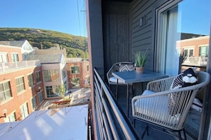 A balcony with a table and two chairs, a potted plant, and a view of buildings and hills.