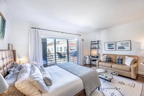 A hotel room with a bed, a sofa, a desk, and a balcony with a view of buildings.