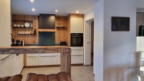 Eine moderne Küche mit Holzausstattung, einer Marmorarbeitsplatte und einem eingebauten Backofen. Im Vordergrund befinden sich zwei Hocker und ein Bild an der Wand.