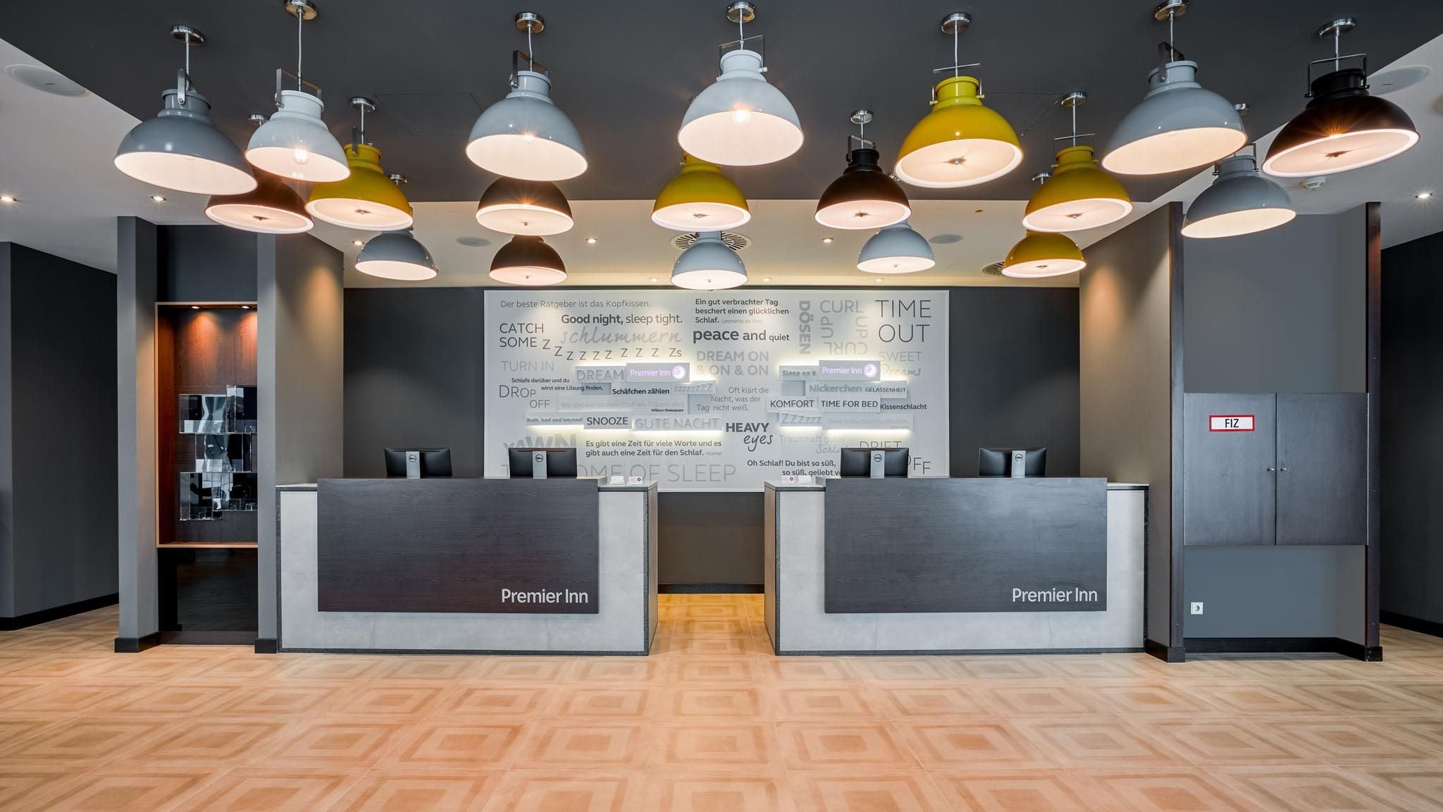 Zona de recepción de un hotel con varias lámparas colgantes, dos mostradores de recepción con la leyenda 'Premier Inn' y una pared grande con varios textos y símbolos.