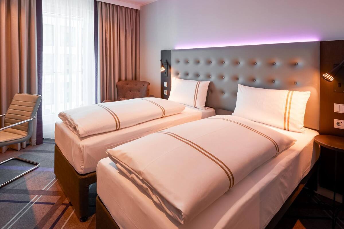 A hotel room with two beds, a chair, a desk, and a lamp.