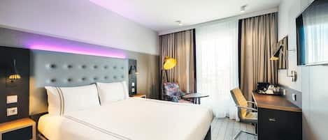 A hotel room with a large bed, a desk, a chair, a lamp, and a TV.