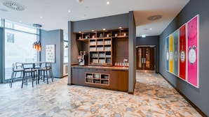 Un bar moderno con estantes llenos de tazas y vasos, un espacio de asientos con mesas y sillas, y una pared colorida con relojes.