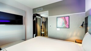 Habitación de hotel con cama, televisor de pantalla plana en la pared, un cuadro y una mesita de noche con lámpara.