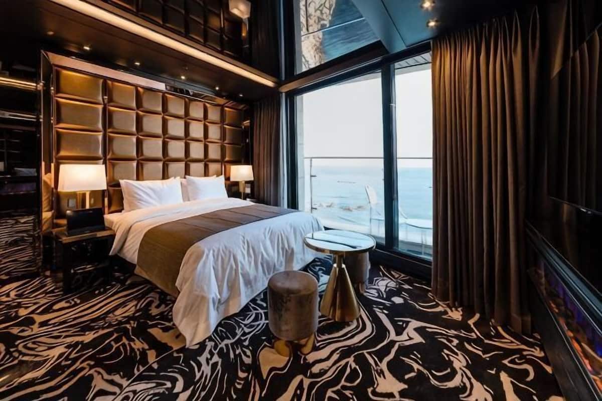 A hotel room with a large bed, a small round table, a view of the sea, and a bookshelf.