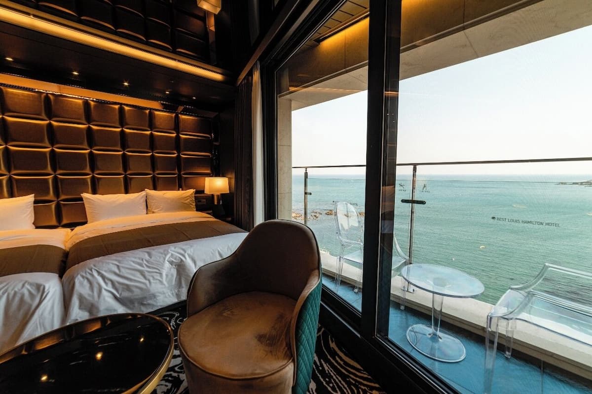 A modern hotel room with a large bed, a seating area with a chair and a table, and a balcony with a view of the sea.