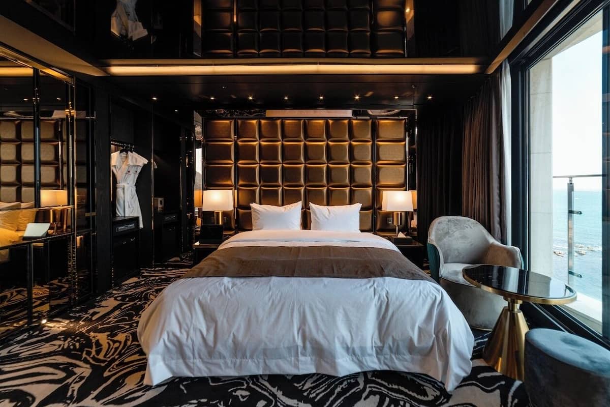 A modern hotel room with a large bed, a seating area with a table, and a view of the sea.