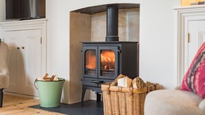A cozy room with a wood-burning stove, a green bucket, and a wicker basket filled with firewood. - West Cottage -  charming seaside cottage by Bloom Stays - 4 guests  in 2 bedrooms (Dungeness)