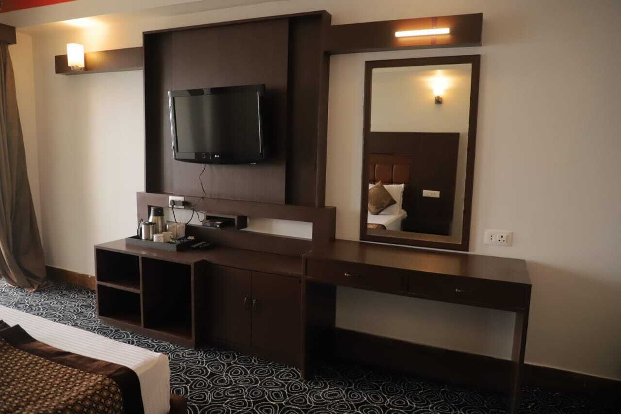 A hotel room with a large flat-screen TV mounted on the wall, a mirror above the console table, and a patterned carpet.